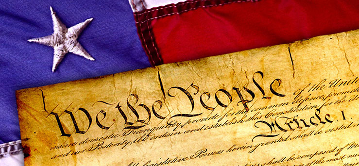 We the People printed on parchment. The parchment is lying on top of the American Flag with a single stitched star appearing in the cropped frame of the image